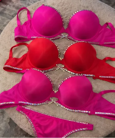 Three luxury satin push-up bras in vibrant magenta pink and scarlet red colors, featuring sparkling rhinestone trim and matching panties, arranged on a beige textured ottoman.